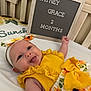 baby, infant, smiling, tongue_out, bed, headband, yellow_clothing, sunflower_pattern, letter_board, text, milestone, cute, happy, portrait, child, indoor, person, fabric, playful, baby_girl