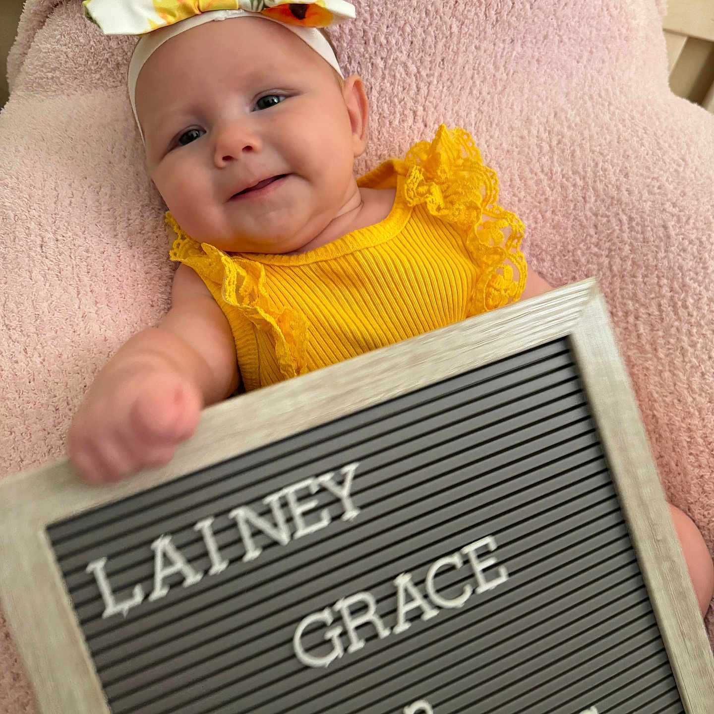 Lainey is registered to the contest to win money with this photo: baby, blanket, child, cute, face, happy, headband, indoors, infant, newborn, person, portrait, resting, sign, skin, smiling, soft_texture, text_board, yellow_clothing, young_child