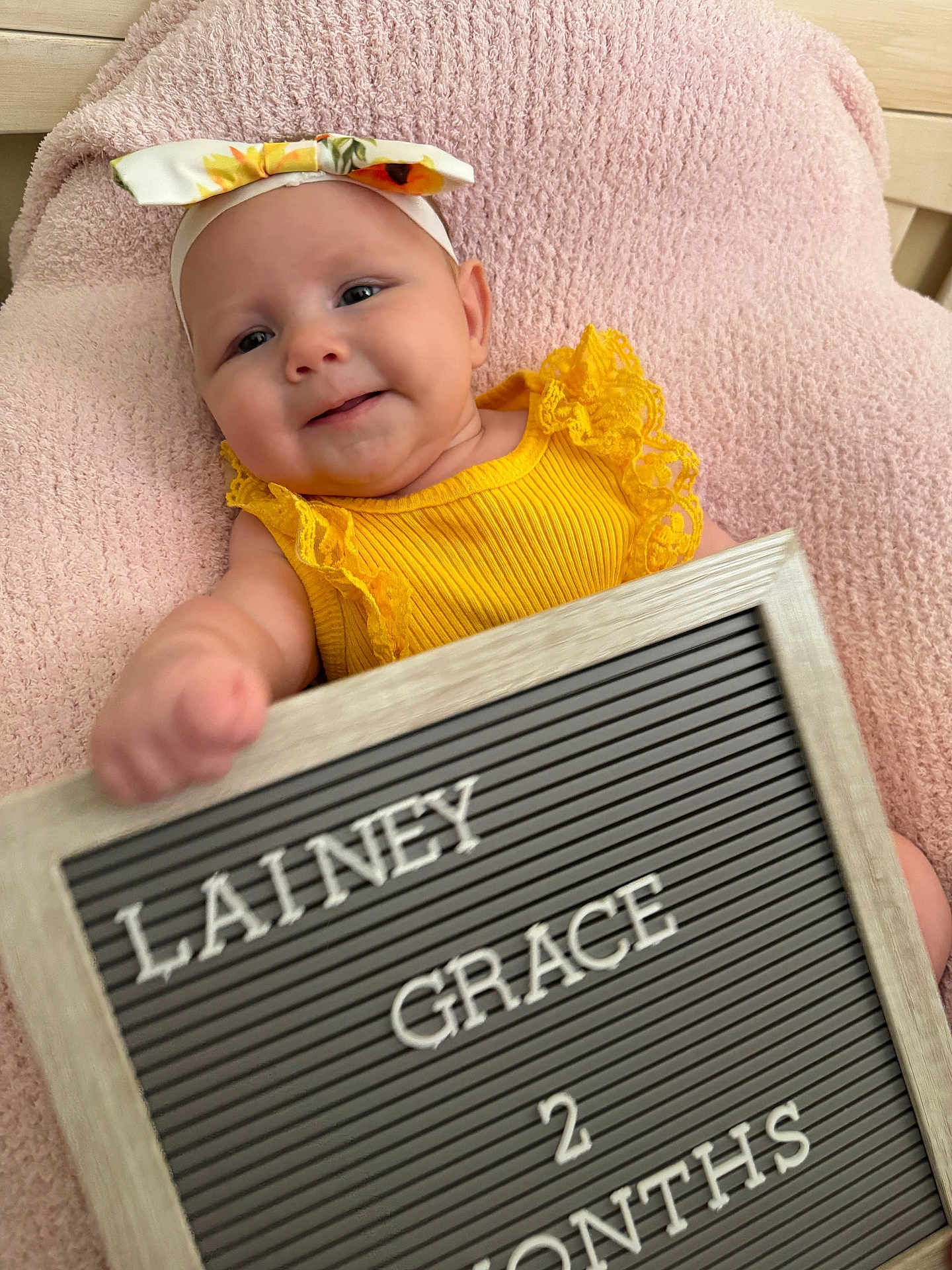 Lainey is registered to the contest to win money with this photo: baby, infant, smiling, yellow_clothing, headband, blanket, sign, text_board, portrait, cute, child, person, indoors, soft_texture, happy, young_child, face, skin, newborn, resting