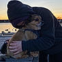 dog, person, hoodie, hug, sunset, water, dock, wooden_plank, leash, harness, hand, face, pet, owner, affection, small_dog, dusk, sky, waterfront, outdoor