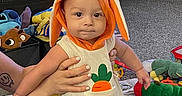 Dash joined the competition — help win amazing prizes! baby, child, orange_outfit, bunny_ears, toys, living_room, carpet, bookshelf, leather_chair, person, hands, plush_toys, colorful, cute, smiling, infant, playmat, socks, tattoos, home