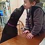 dog, child, boy, indoor, table, wooden_table, affection, pet, black_dog, purple_collar, smile, kitchen, chair, home, interaction, friendship, cute, happy, face, human