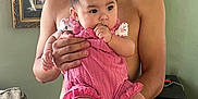 Arya is registered to the contest to win money with this photo: baby, child, man, holding, pink_clothing, bow, socks, indoor, portrait, curly_hair, jeans, furniture, couch, picture_frame, green_wall, hand, face, skin, person, home