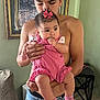 baby, child, man, holding, pink_clothing, bow, socks, indoor, portrait, curly_hair, jeans, furniture, couch, picture_frame, green_wall, hand, face, skin, person, home