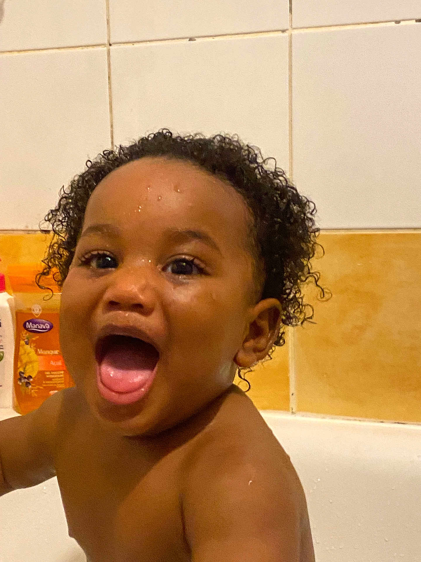 Shëlly a rejoint le concours — aidez-le/la à gagner de superbes lots ! child, toddler, bathtub, bathroom, water, curly_hair, happy, smiling, wet_skin, face, mouth_open, person, tile_wall, soap_bottle, orange_bottle, white_bottle, splashing, skin, head, expression