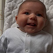 Luna is registered to the contest to win money with this photo: baby, infant, smile, face, white_clothing, buttoned_outfit, lying_down, soft_mat, cheeks, happy, cute, closeup, indoors, child, young, head, person, expression, skin, button