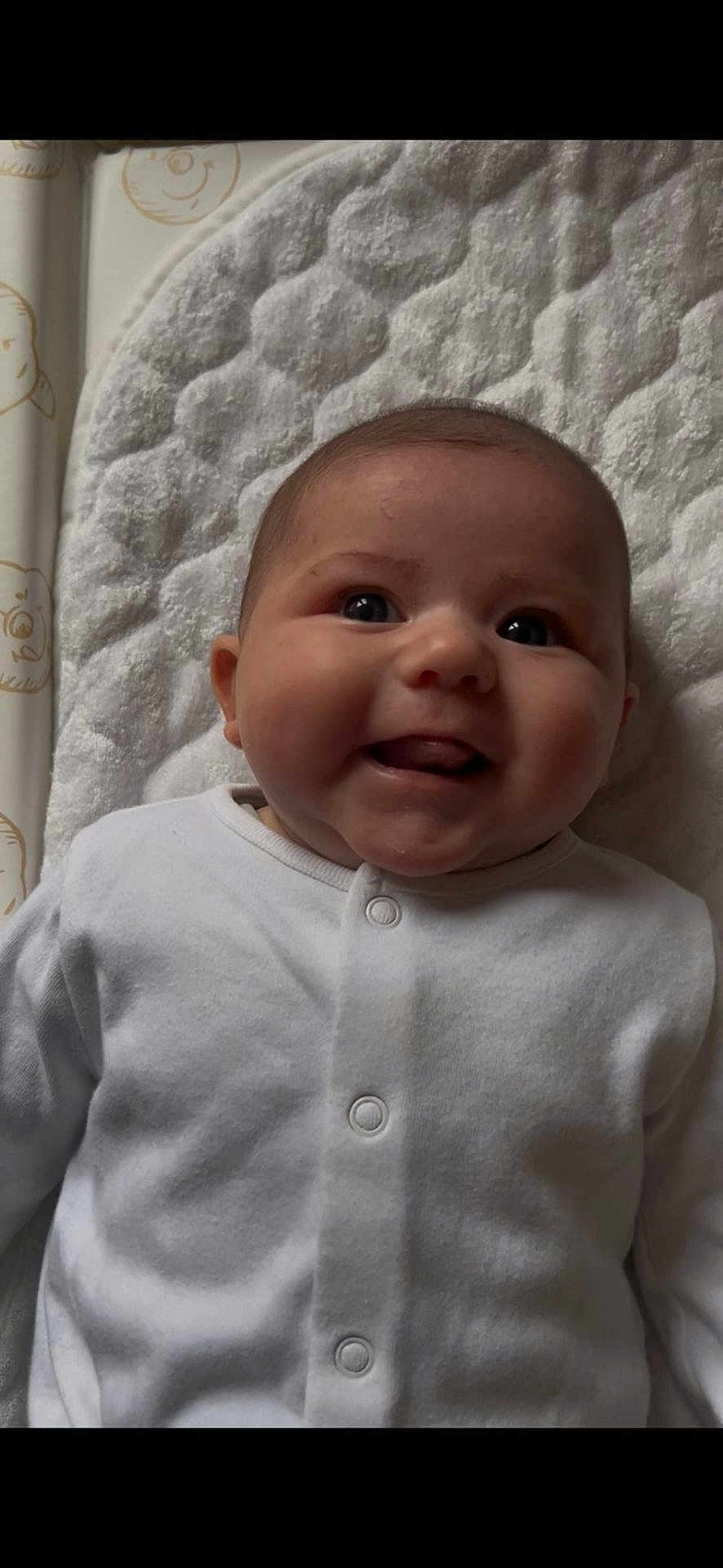 Luna is registered to the contest to win money with this photo: baby, infant, smile, face, white_clothing, buttoned_outfit, lying_down, soft_mat, cheeks, happy, cute, closeup, indoors, child, young, head, person, expression, skin, button