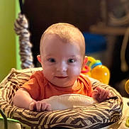 Buck is registered to the contest to win money with this photo: baby, baby_seat, child, closeup, colorful, cute, face, happy, head, indoor, infant, orange_clothing, person, playtime, seated, smiling, soft_light, toy, young, zebra_pattern