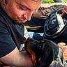 man, dog, rottweiler, car, steering_wheel, tattoo, arm, black_shirt, sunlight, window, closeup, pet, companion, affection, interaction, portrait, indoor, animal, human, friendship