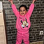child, toddler, pink_clothing, minnie_mouse, sweatshirt, pants, white_shoes, raised_arms, smiling, indoor, brick_wall, wooden_floor, happy, cute, playful, person, fashion, casual, portrait, standing