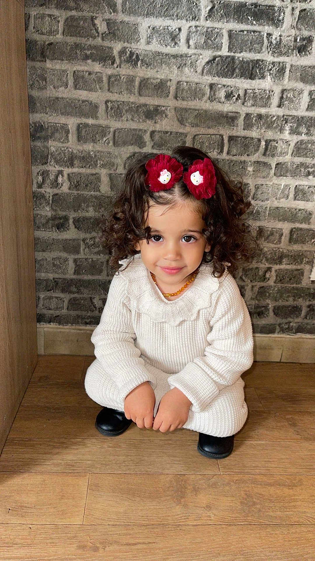 Eyline a rejoint le concours — aidez-le/la à gagner de superbes lots ! child, curly_hair, hair_clips, red_flower, white_knit_outfit, black_shoes, smile, squatting, wooden_floor, brick_wall, indoor, cute, portrait, young_child, fashion, accessories, happy, cozy, casual, person