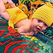Nate a rejoint le concours — aidez-le/la à gagner de superbes lots ! baby, child, knitted_hat, yellow_clothing, blanket, cartoon_character, smiling, lying_down, indoor, cute, toddler, face, person, colorful, soft_texture, plush_toy, happy, portrait, cozy, playful