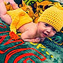 baby, child, knitted_hat, yellow_clothing, blanket, cartoon_character, smiling, lying_down, indoor, cute, toddler, face, person, colorful, soft_texture, plush_toy, happy, portrait, cozy, playful