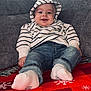 baby, child, smiling, striped_hoodie, denim_jeans, socks, couch, red_blanket, snowflake_pattern, indoors, happy, drool, casual_clothing, cozy, portrait, seated, cute, young, infant, relaxed