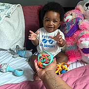 Javaree is registered to the contest to win money with this photo: baby, child, smiling, toy, hand, bed, pillow, stuffed_animal, plush, clothing, indoor, person, happy, infant, cute, playful, toddler, african_american, blanket, tattoo
