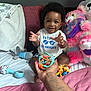 baby, child, smiling, toy, hand, bed, pillow, stuffed_animal, plush, clothing, indoor, person, happy, infant, cute, playful, toddler, african_american, blanket, tattoo