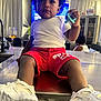 Javaree is registered to the contest to win money with this photo: toddler, child, indoor, shorts, white_shirt, sneakers, pacifier, table, kitchen_counter, curtains, cabinet, living_room, glass, person, hair, furniture, toy, casual_clothing, seated, curious