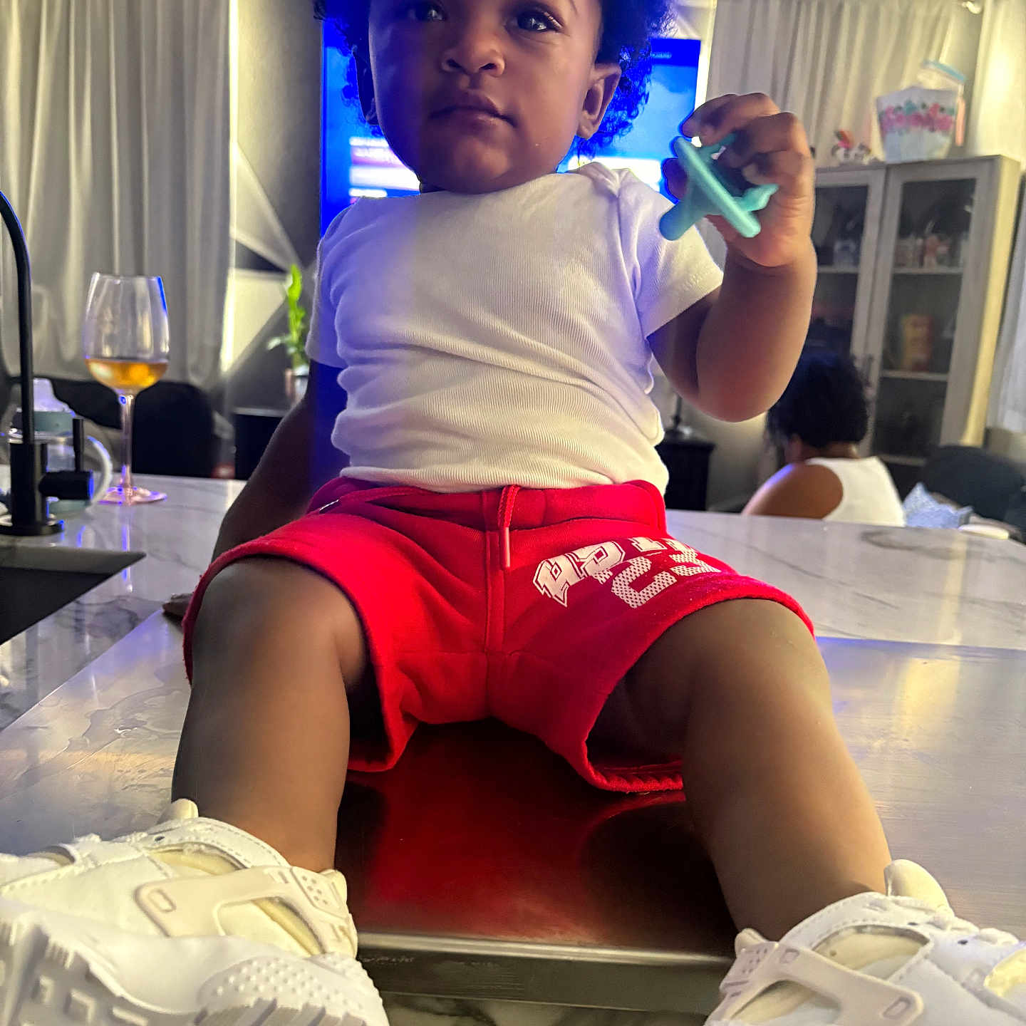 Javaree is registered to the contest to win money with this photo: cabinet, casual_clothing, child, curious, curtains, furniture, glass, hair, indoor, kitchen_counter, living_room, pacifier, person, seated, shorts, sneakers, table, toddler, toy, white_shirt