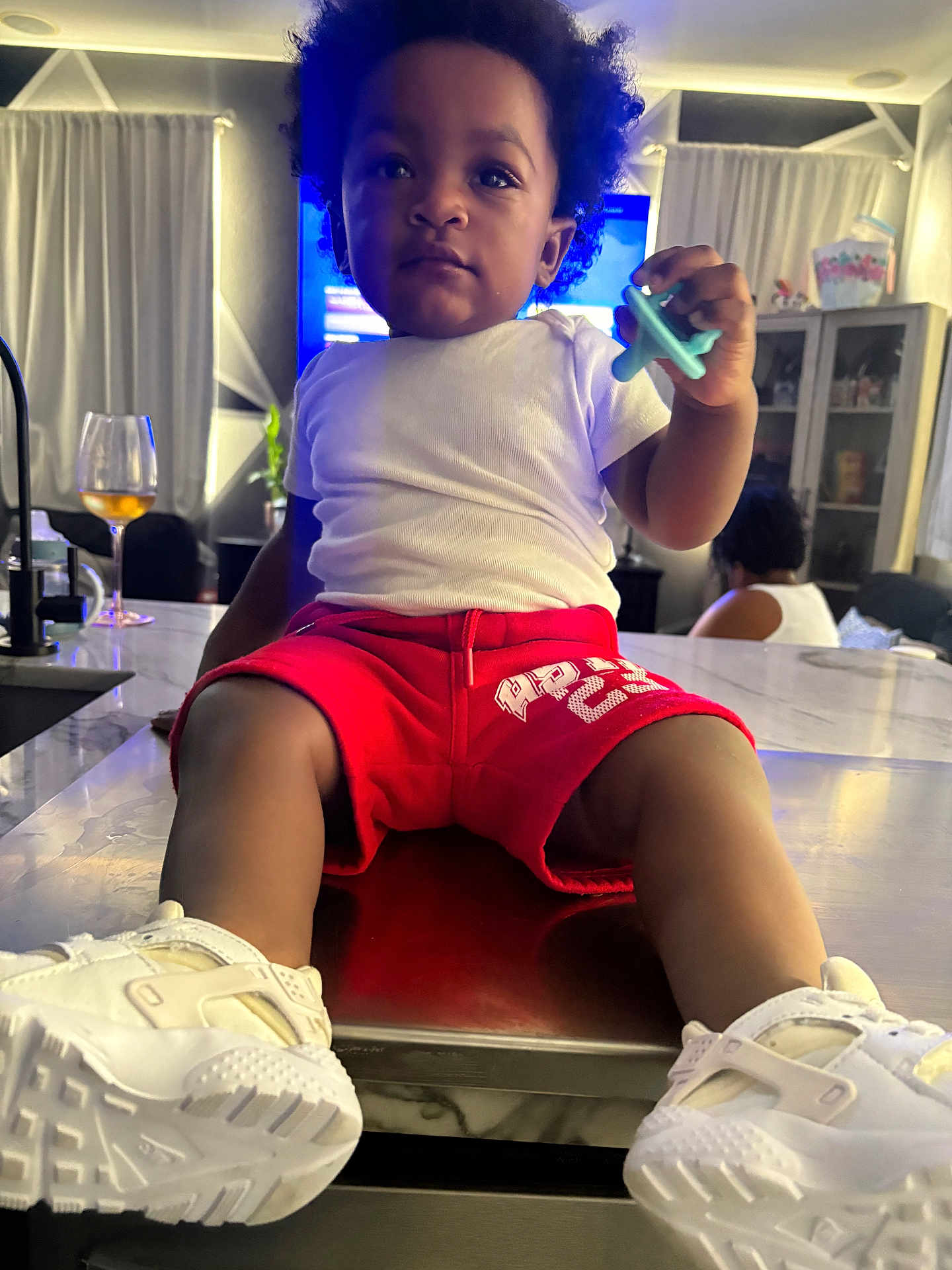 Javaree is registered to the contest to win money with this photo: toddler, child, indoor, shorts, white_shirt, sneakers, pacifier, table, kitchen_counter, curtains, cabinet, living_room, glass, person, hair, furniture, toy, casual_clothing, seated, curious