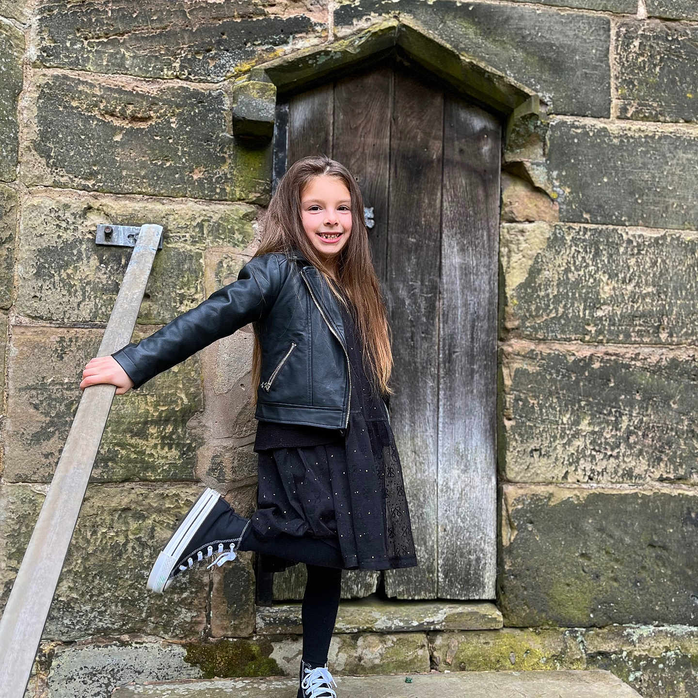 Raluca joined the competition — help win amazing prizes! black_dress, black_jacket, casual_clothing, child, daylight, fashion, girl, happy, long_hair, outdoor, playful, portrait, railing, smile, sneakers, standing, steps, stone_wall, wooden_door, young_person