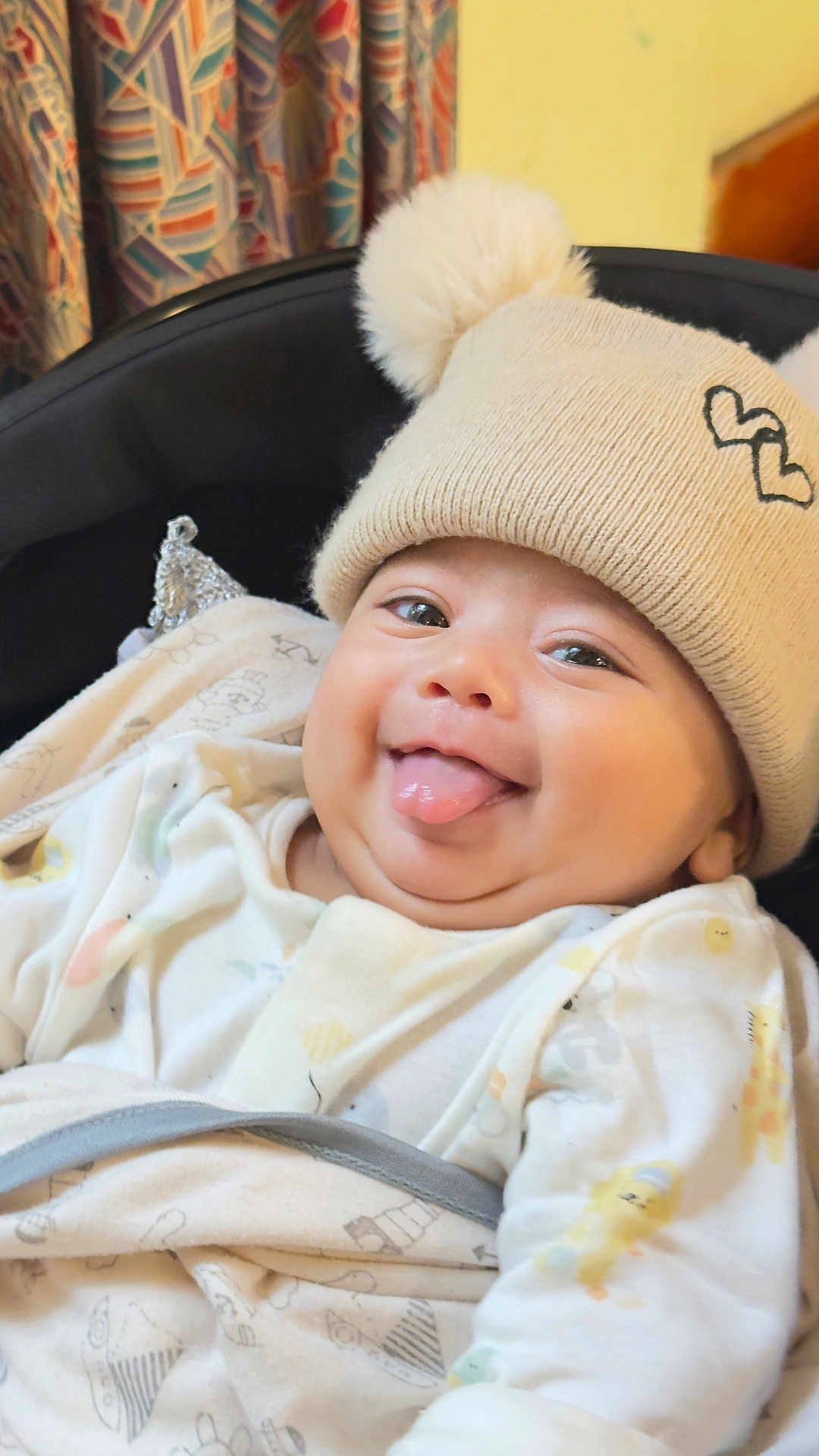 Ammar is registered to the contest to win money with this photo: baby, child, tongue_out, smiling, hat, pom_poms, beige_hat, cozy_clothing, patterned_clothes, indoor, curtain, seat, happy, cute, closeup, infant, playful, face, person, portrait