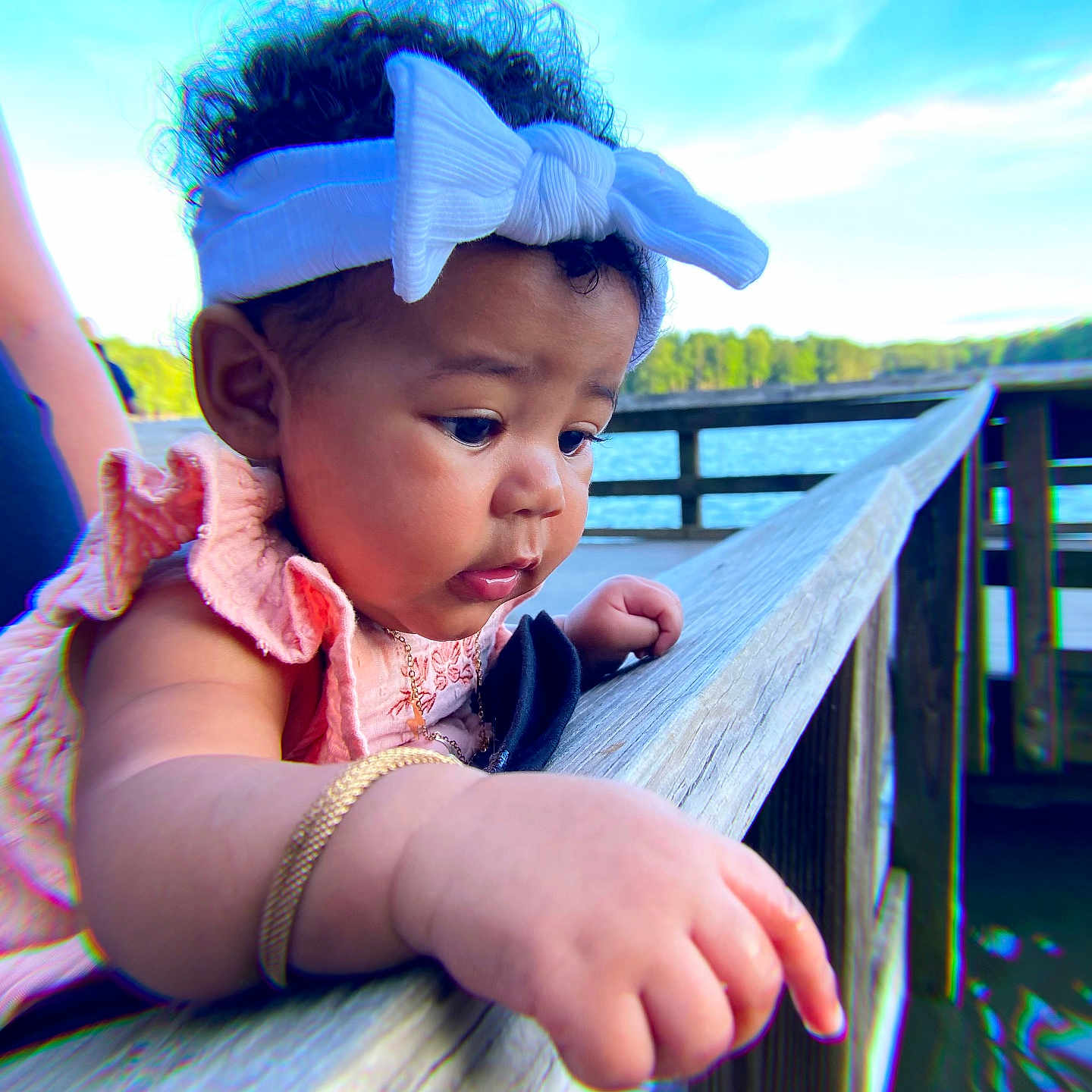 Ramiya is registered to the contest to win money with this photo: arm, baby, bow, bracelet, child, closeup, curly_hair, daylight, hand, headband, lake, nature, outdoor, person, pink_clothing, portrait, sky, trees, water, wooden_railing
