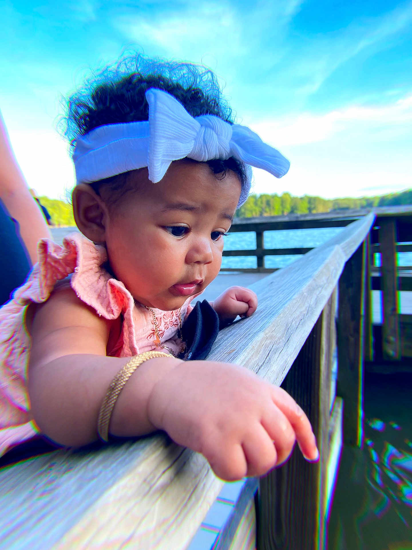 Ramiya is registered to the contest to win money with this photo: baby, child, headband, bow, curly_hair, arm, hand, wooden_railing, lake, water, outdoor, nature, trees, sky, daylight, portrait, closeup, pink_clothing, bracelet, person