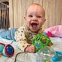 baby, child, smiling, toy, play_mat, indoor, happy, infant, clothing, hand, face, wooden_wall, colorful, background, playtime, cute, person, blue_pants, green_toy, pink_blanket