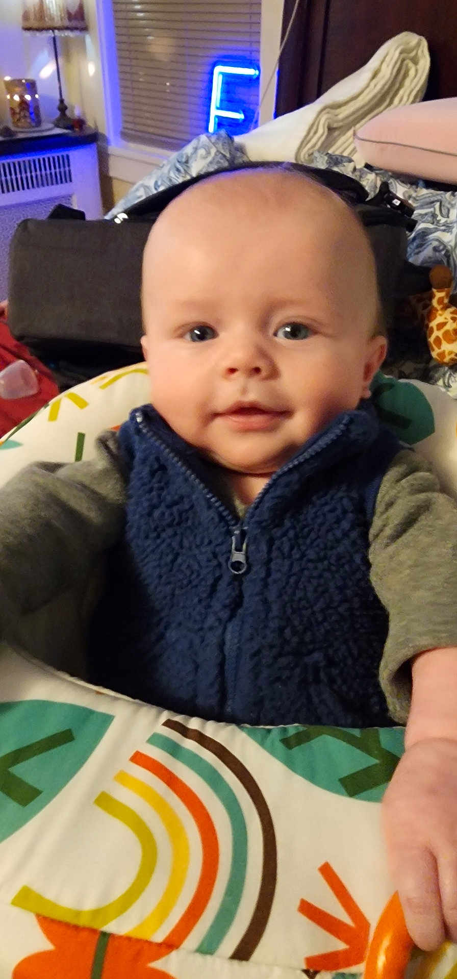 Jameson joined the competition — help win amazing prizes! baby, child, infant, smile, face, blue_vest, fleece, baby_seat, toy, giraffe, hand, indoor, lamp, neon_light, window, curtain, blanket, colorful, cozy, portrait