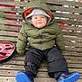 baby, child, infant, smiling, hooded_jacket, green_jacket, puffy_jacket, blue_boots, rain_boots, black_pants, wooden_bench, bench_slats, outdoor, winter_clothing, sitting, portrait, happy, rosy_cheeks, toy_disc, cold_weather
