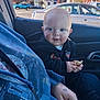 Dominik is registered to the contest to win money with this photo: baby, infant, child, car_interior, car_window, parking_lot, cookie, crumbs, hand, jacket, car_door, seat, tattoo, heart_tattoo, smile, cheeks, daylight, storefront, trees, passenger