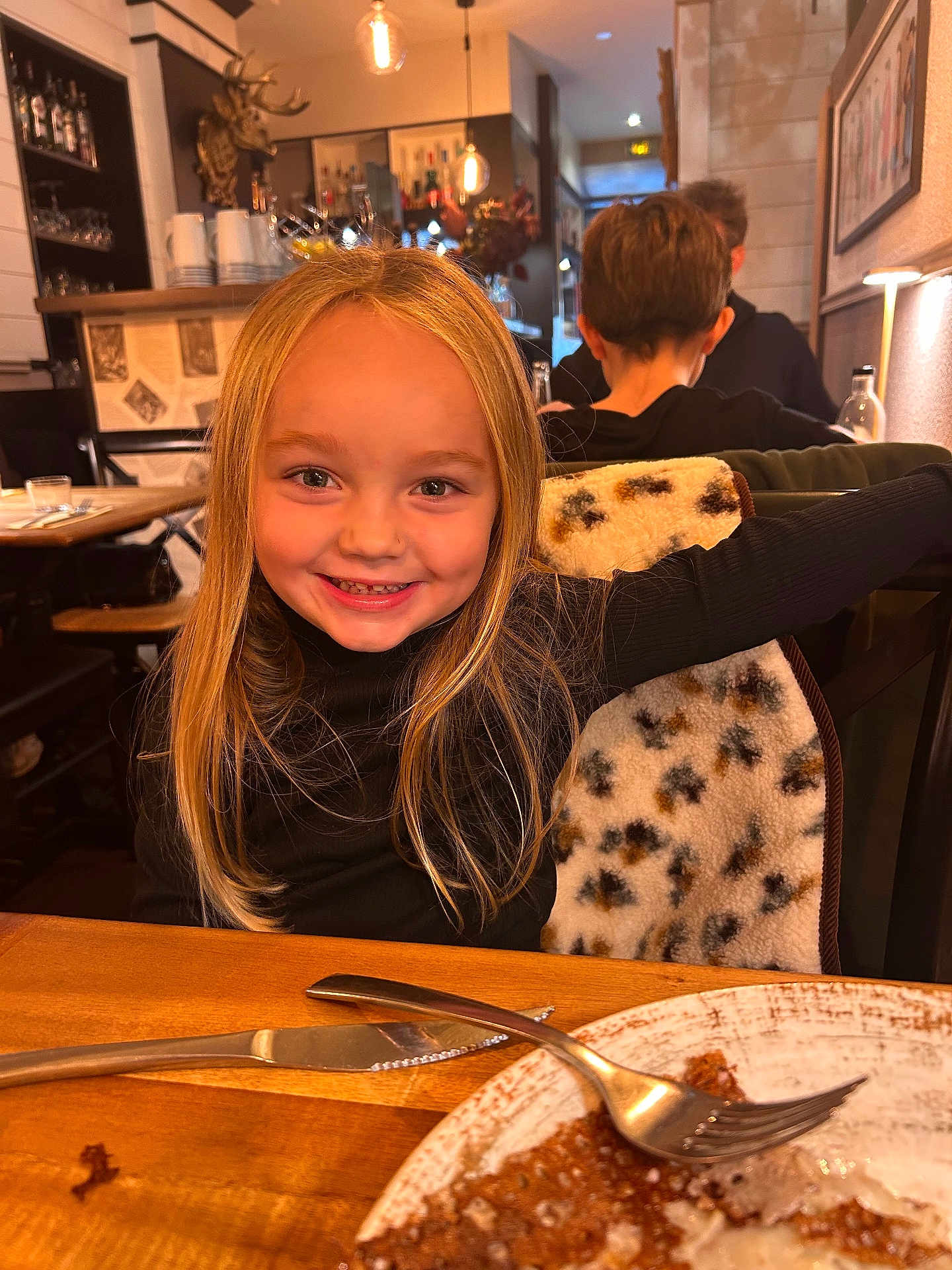 Hina participe au concours pour gagner de l'argent avec cette photo : child, smiling, blonde_hair, restaurant, table, plate, fork, knife, food, cozy, indoor, dining, happy, person, long_hair, warm_lighting, wooden_table, background_people, chair, jacket