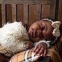 Kayna a rejoint le concours — aidez-le/la à gagner de superbes lots ! baby, sleeping, infant, wooden_chair, plaid_pillow, headband, bow, fluffy_clothing, hand, peaceful, closeup, portrait, soft_texture, brown_background, cozy, child, cute, relaxed, indoor, resting