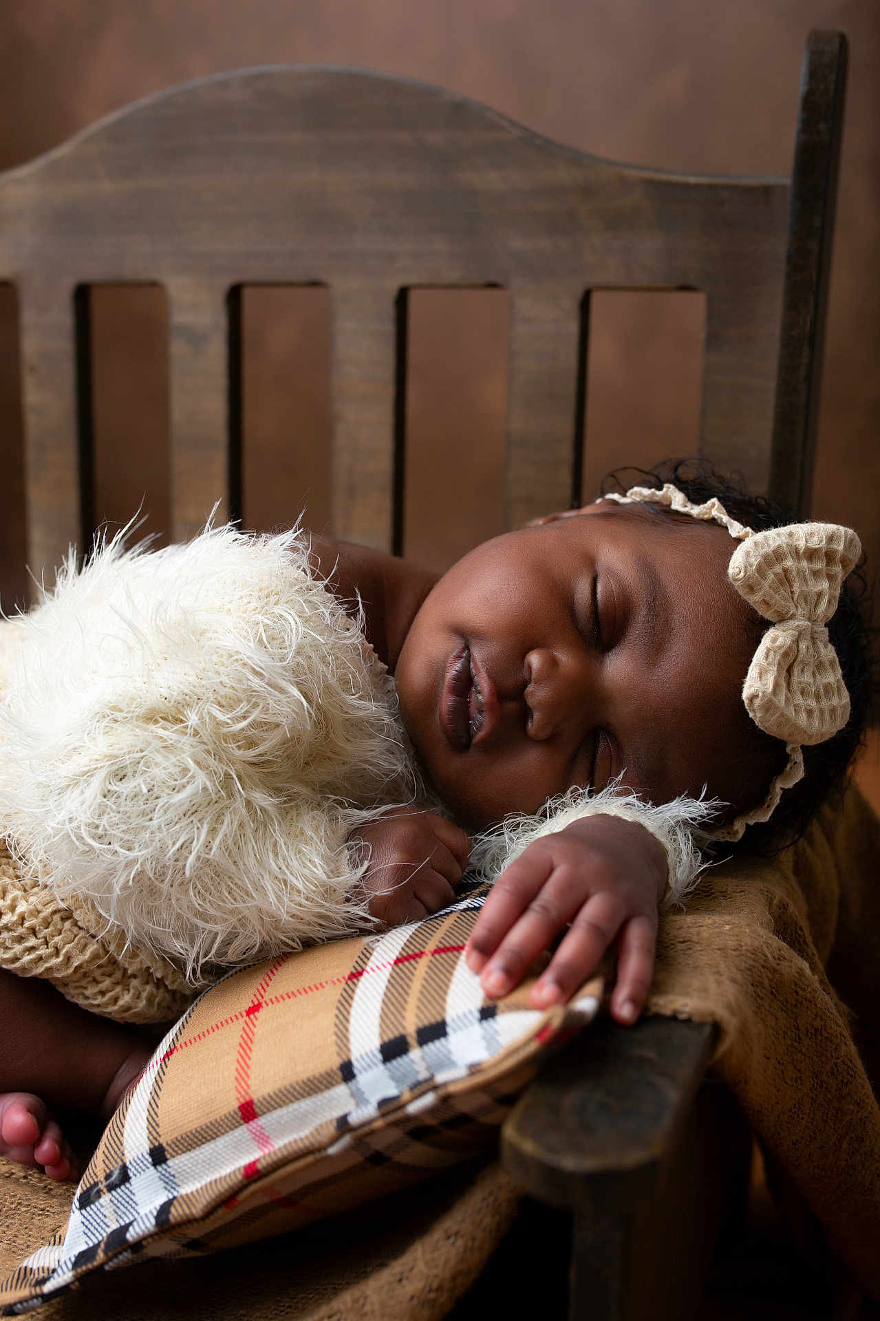 Kayna a rejoint le concours — aidez-le/la à gagner de superbes lots ! baby, sleeping, infant, wooden_chair, plaid_pillow, headband, bow, fluffy_clothing, hand, peaceful, closeup, portrait, soft_texture, brown_background, cozy, child, cute, relaxed, indoor, resting