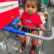 Alaiah joined the competition — help win amazing prizes! black_hair, blanket, box, child, clothing, curly_hair, eyes, face, floor, grocery_store, hand, indoor, person, red_bow, red_shirt, retail, shopping, shopping_cart, sneakers, toddler