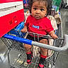 black_hair, blanket, box, child, clothing, curly_hair, eyes, face, floor, grocery_store, hand, indoor, person, red_bow, red_shirt, retail, shopping, shopping_cart, sneakers, toddler