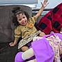 toddler, child, smiling, curly_hair, couch, indoor, toy, doll, purple, plaid_skirt, bear_patch, waving, happy, person, clothing, window_blind, blanket, red_black, hair_bun, playful