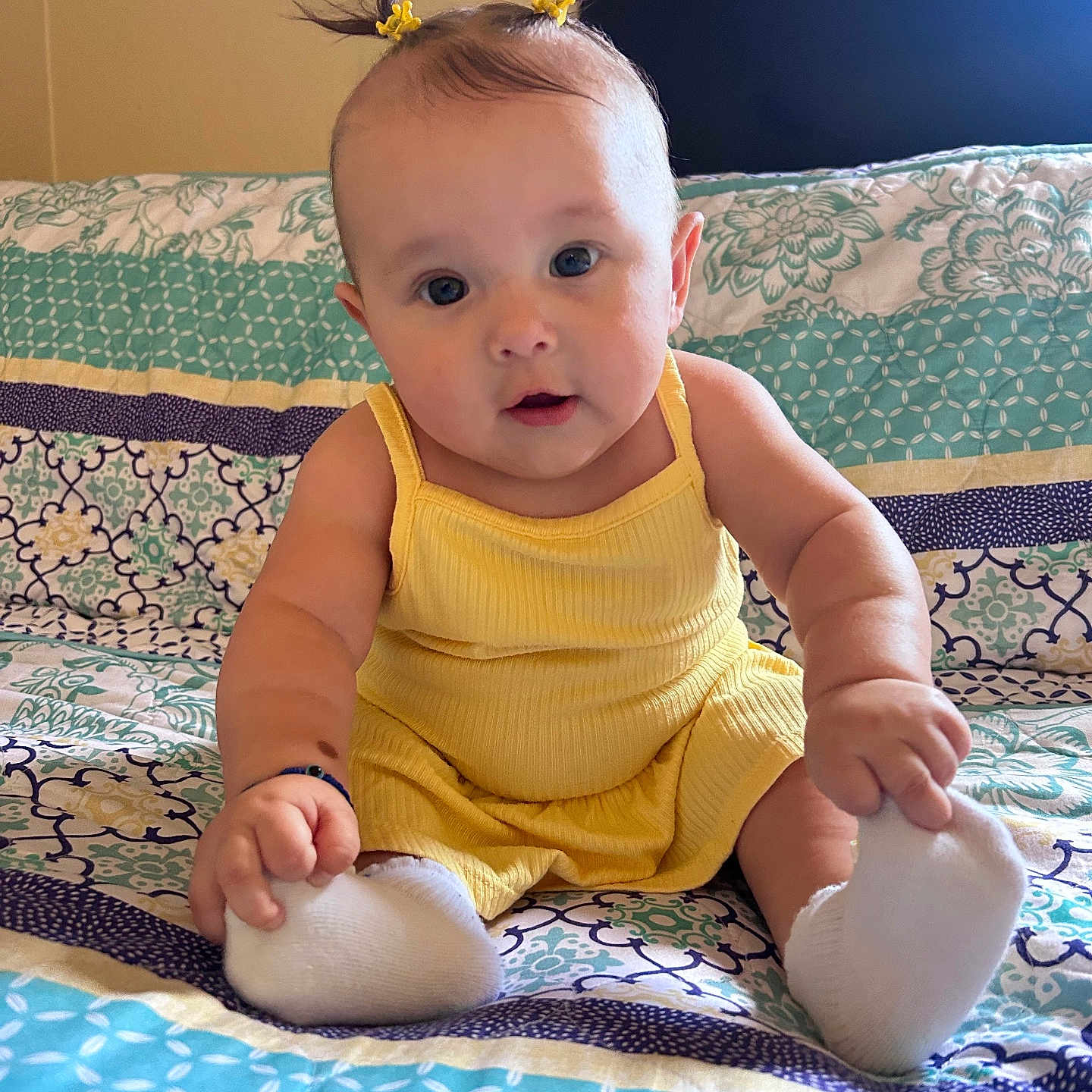 Maricruz joined the competition — help win amazing prizes! baby, bedspread, child, curious, cute, expression, face, feet, hand, indoor, infant, patterned_bedspread, person, pony_tail, portrait, sitting, skin, soft_lighting, white_socks, yellow_outfit