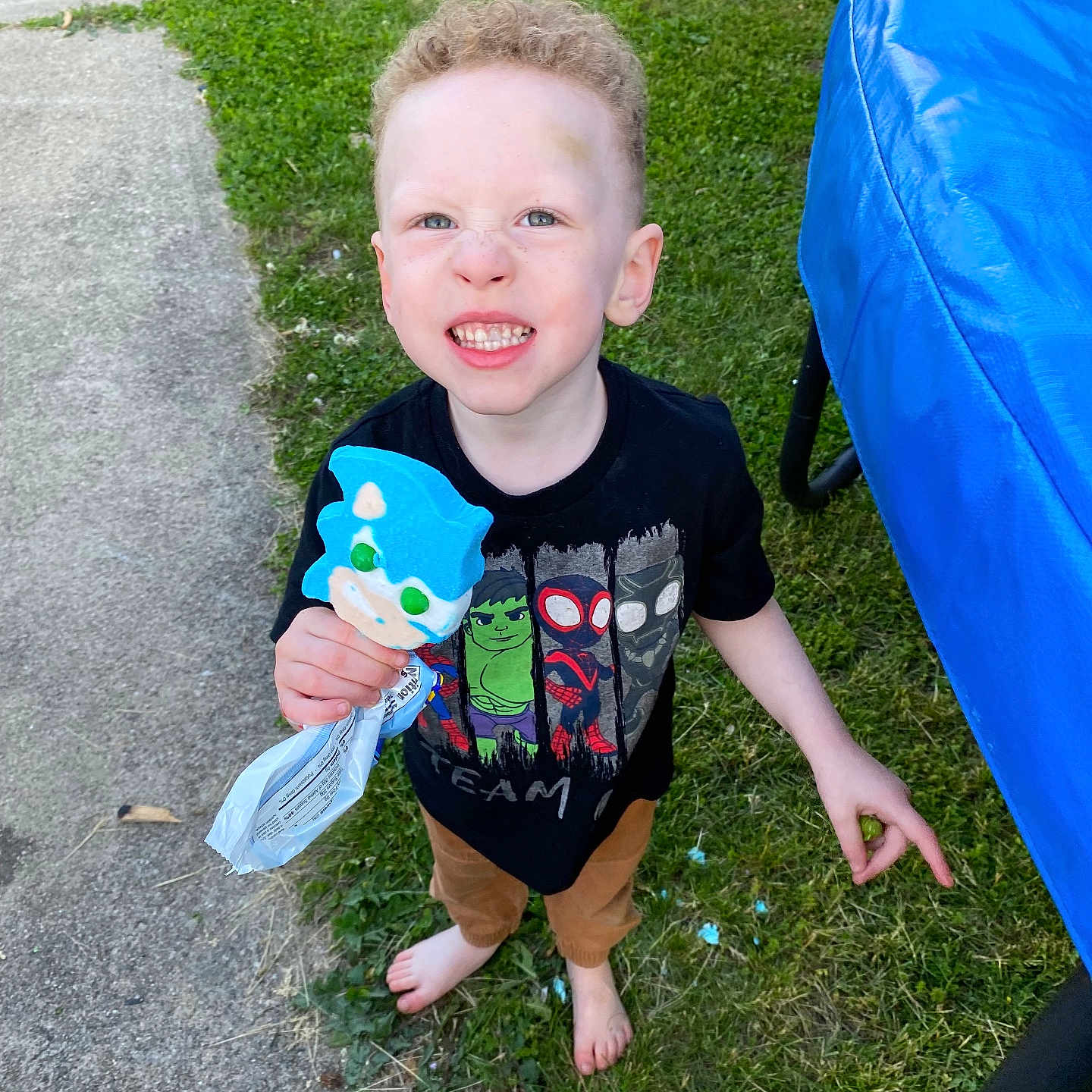 Cortez joined the competition — help win amazing prizes! barefoot, black_tshirt, blue_cookie, boy, brown_pants, casual_clothing, child, curly_hair, daylight, fun, grass, outdoor, playful_expression, pointing, sidewalk, smiling, standing, superhero_cookie, trampoline, young