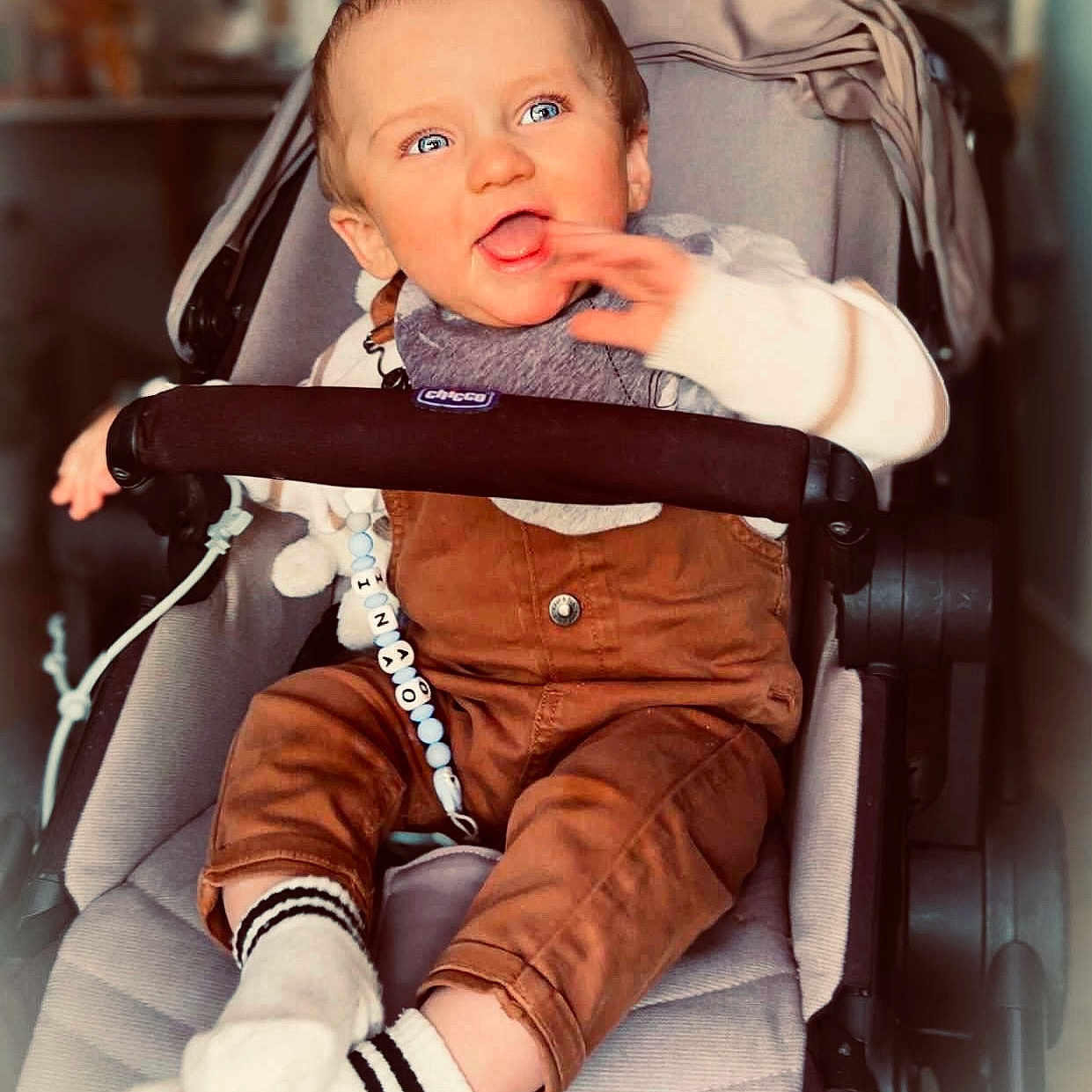 Inao participe au concours pour gagner de l'argent avec cette photo : baby, blue_eyes, brown_overalls, child, clothing, comfort, cute, hand, happy, home, indoor, infant, person, portrait, seat, smiling, socks, stroller, toy, young