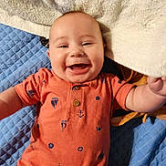 River is registered to the contest to win money with this photo: baby, smiling, infant, orange_outfit, blue_quilt, towel, happy, face, cute, child, lying_down, home, apparel, patterned_clothing, cheerful, skin, portrait, person, indoor, playful