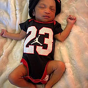 Terrion is registered to the contest to win money with this photo: baby, sleeping, infant, onesie, black_clothing, red_trim, number_23, beanie, hat, blanket, soft_texture, cozy, indoor, resting, cute, peaceful, small, child, portrait, human