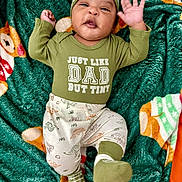 Demetrius Jr joined the competition — help win amazing prizes! baby, infant, child, green_clothing, dinosaur_print, blanket, plush_blanket, smiling, waving_hand, hat, cute, lying_down, indoors, cozy, footwear, patterned_pants, text_on_clothing, happy, portrait, person