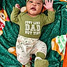 baby, infant, child, green_clothing, dinosaur_print, blanket, plush_blanket, smiling, waving_hand, hat, cute, lying_down, indoors, cozy, footwear, patterned_pants, text_on_clothing, happy, portrait, person