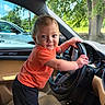 automobile, baby, black_shorts, car, child, dashboard, daylight, greenery, indoor, orange_shirt, parked_car, portrait, seat, smiling, standing, steering_wheel, toddler, trees, vehicle, window