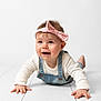 Elizabeth is registered to the contest to win money with this photo: baby, child, clothing, crawling, curious, cute, denim_overalls, eyes, face, hands, happy, headband, indoor, infant, person, pink_bow, portrait, smiling, white_background, white_floor