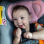 baby, car_seat, smiling, orange_clothing, safety_harness, infant, child, seatbelt, cute, happy, portrait, indoor, warning_label, human, skin, fingers, head, eyes, mouth, hair