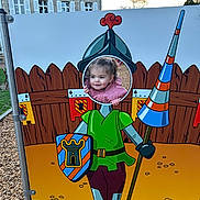 Julianna participe au concours pour gagner de l'argent avec cette photo : building, cartoon, casual_clothing, child, curious, cutout, daylight, face, fence, fun, imagination, knight, lance, outdoor, pink_jacket, playground, shield, shoes, toddler, wood_chips