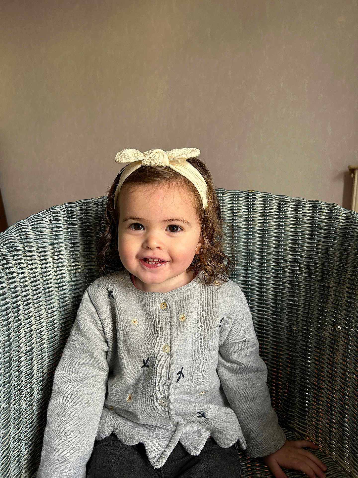 Julianna participe au concours pour gagner de l'argent avec cette photo : toddler, child, smiling, headband, curly_hair, cardigan, gray_clothing, sitting, wicker_chair, indoor, person, happy, cute, portrait, young_child, fashion, cozy, casual, female, relaxed