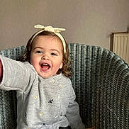 Julianna a rejoint le concours — aidez-le/la à gagner de superbes lots ! toddler, child, smiling, happy, curly_hair, headband, sweater, embroidered, chair, woven_chair, indoor, person, portrait, cute, baby, hand, seated, cozy, warm_clothing, playful